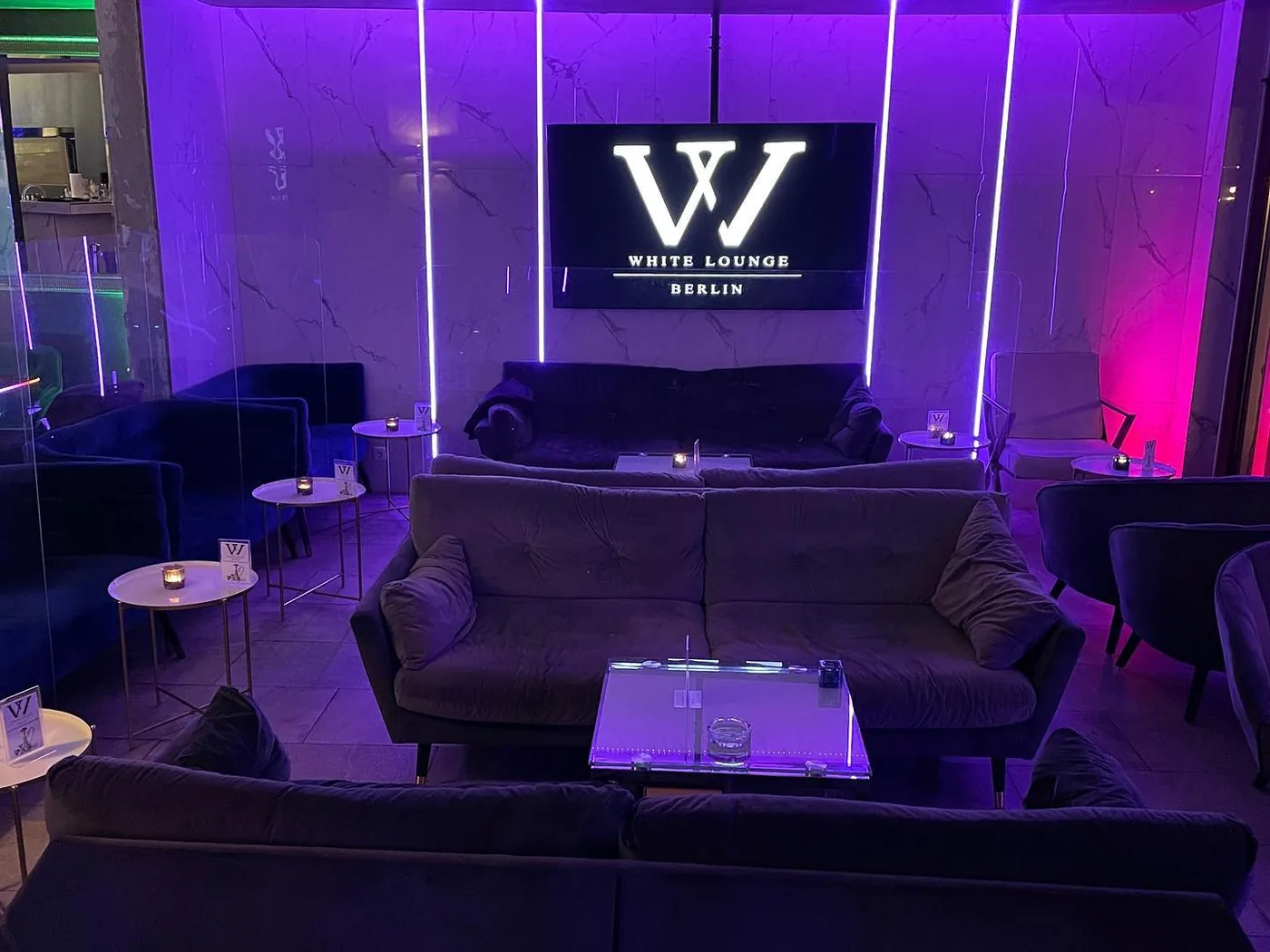 Sofás frente a un gran televisor de pared con el logotipo de White Lounge, entre tiras de luz violeta